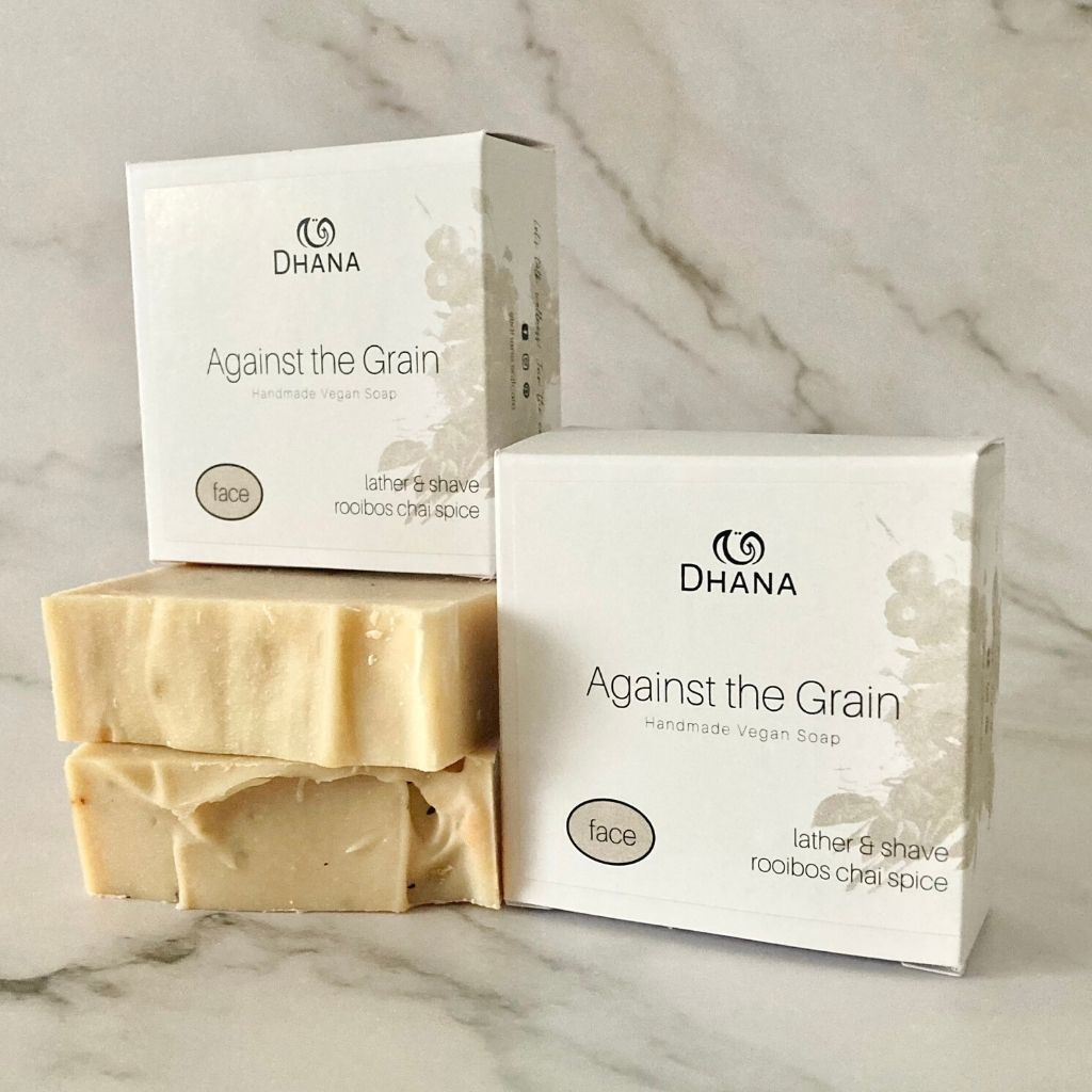 brown bars of soap with boxes on top and beside. The boxes read Against the Grain Shaving Soap from Dhana Selfcare. All on a Marble background.