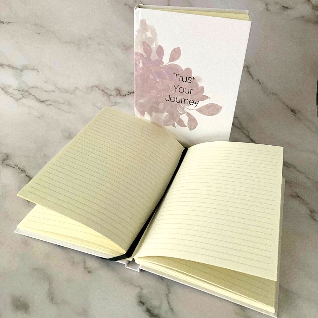 Journal with soft pink botanicals upright and one open with cream lined pages and black bookmark ribbon