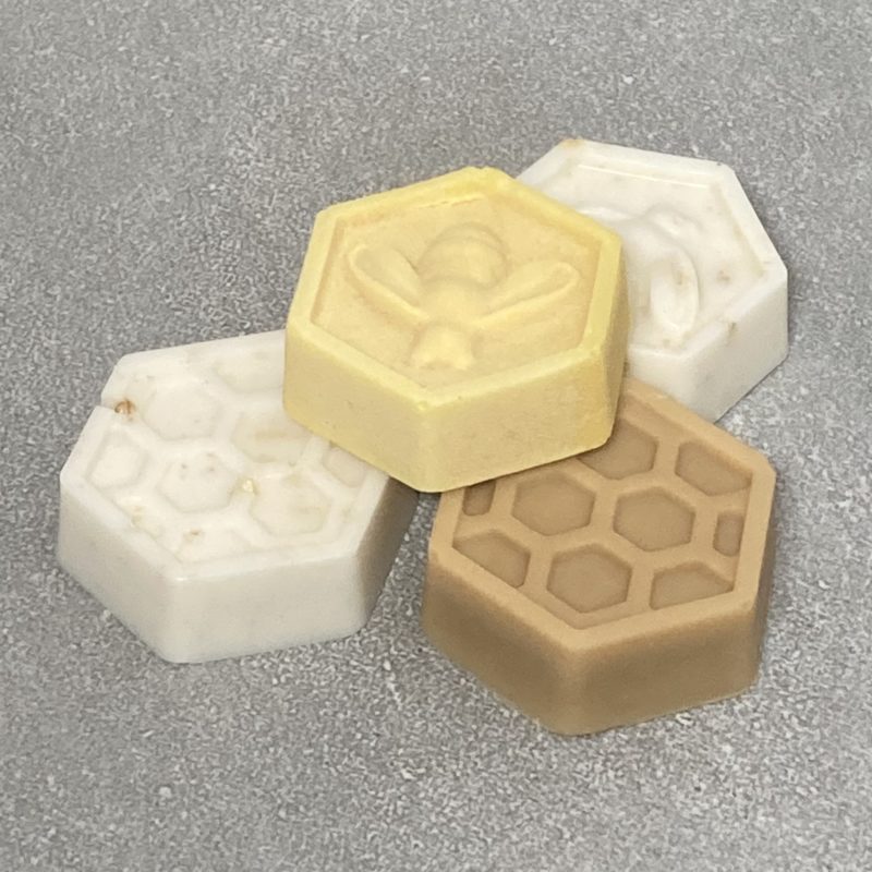 four honeycomb shaped soaps on a grey tile surface. Two are white with oats, one yellow with a bee on the top and another is a deep honey colour with honeycomb texture on top.