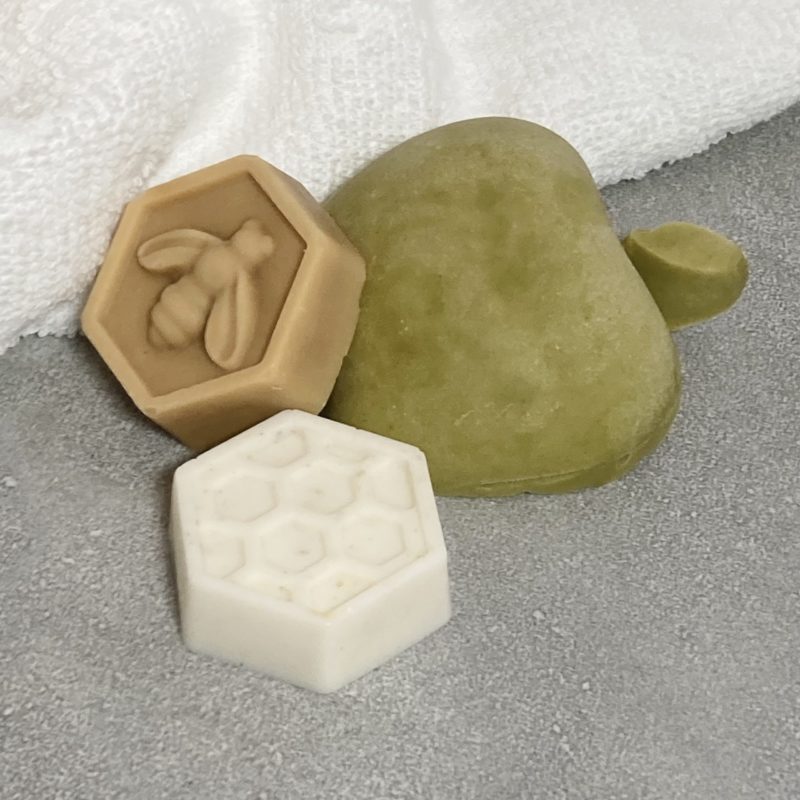on a grey tile sits a green half apple shaped soap, flat side down and two honeycomb shaped soaps. One is white with honeycomb design and the other is brown like honey with a bee on it. These make up the Apple & Honey Rosh Hashanah Gift Collection.