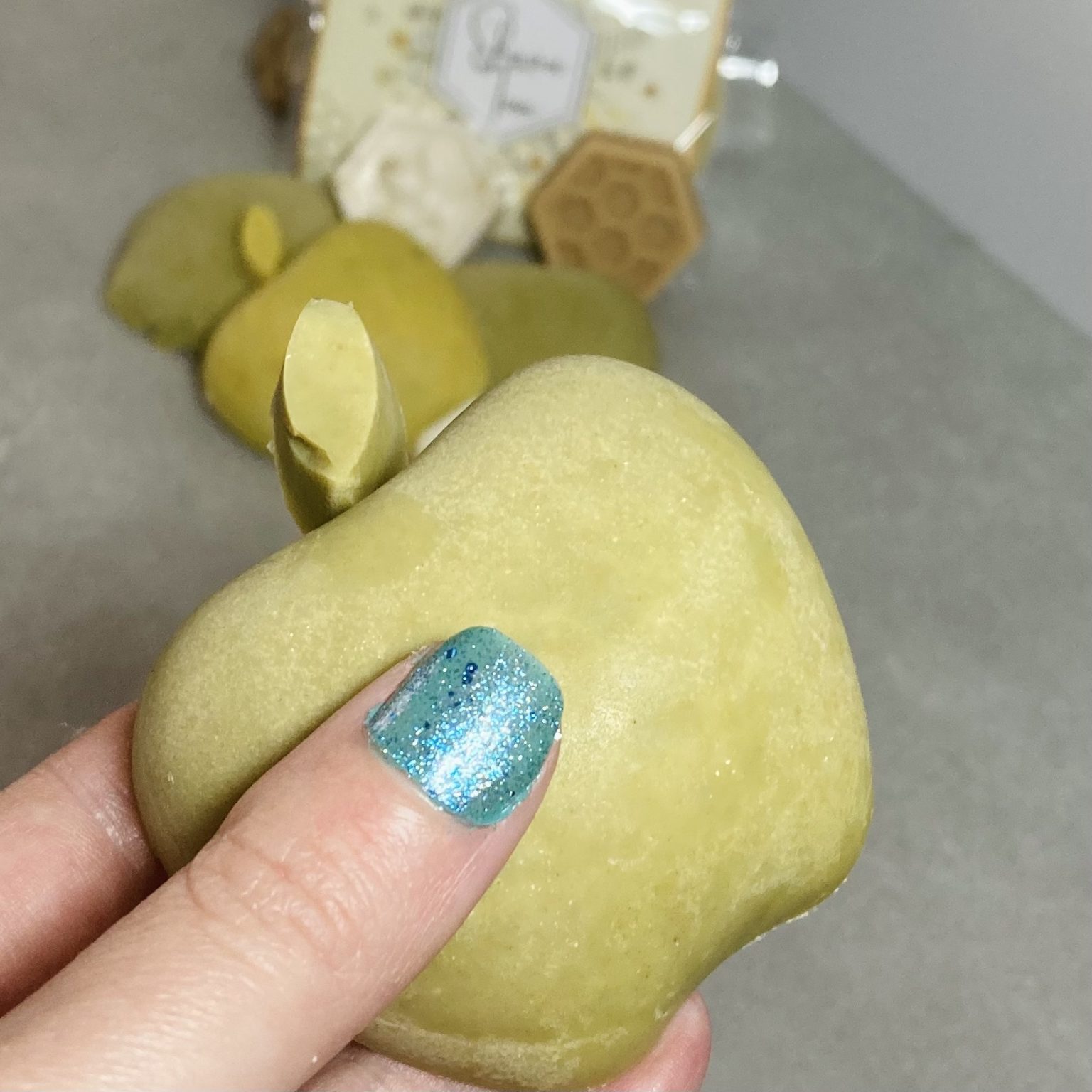 a hand with turquoise finger nail holding a green apple soap with other honey and apple soaps in the background. They are out of focus. The held bar of soap is just wider than three fingers.