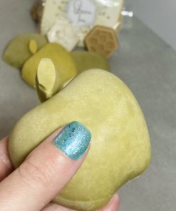 a hand with turquoise finger nail holding a green apple soap with other honey and apple soaps in the background. They are out of focus. The held bar of soap is just wider than three fingers.