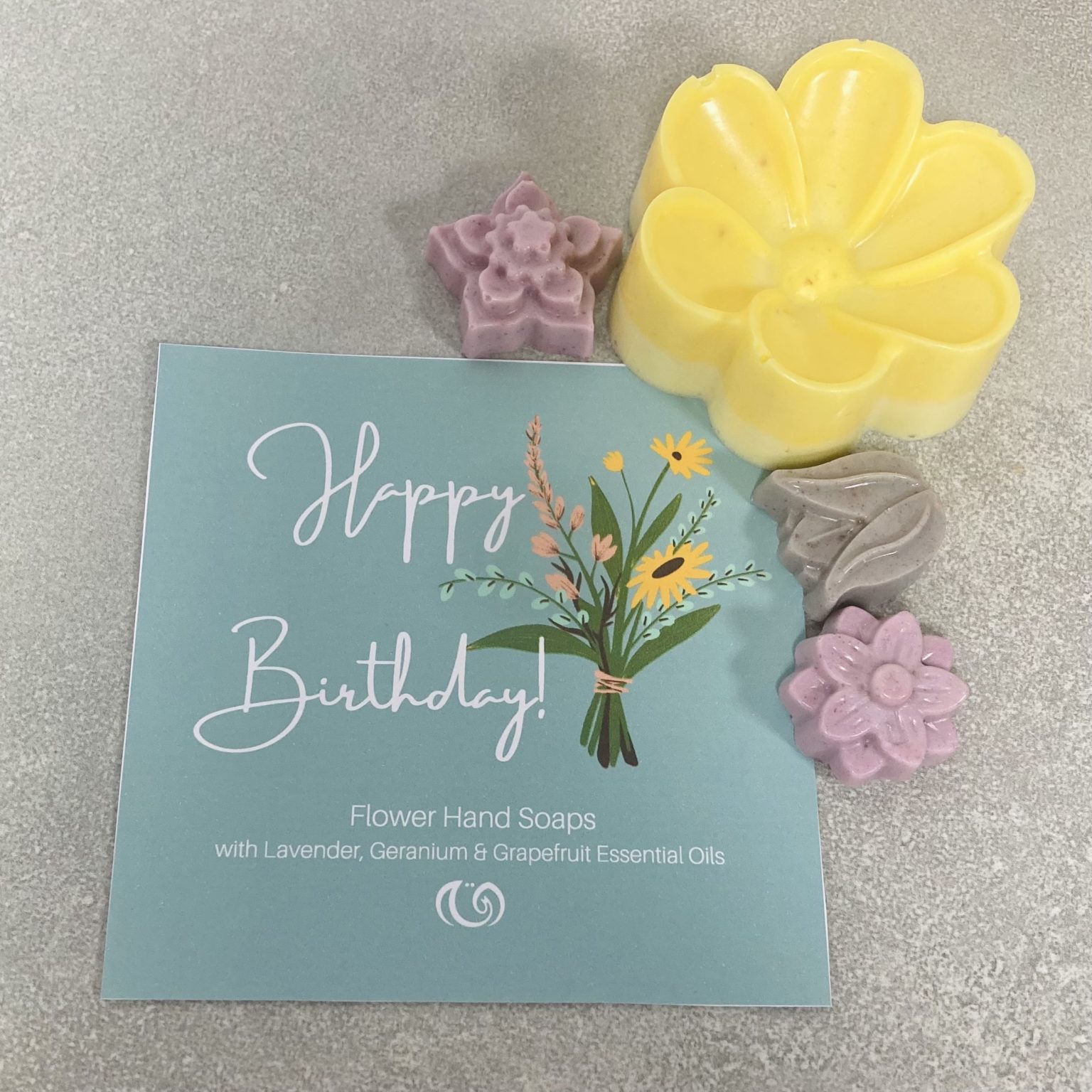 2 small purple a small grey and big yellow soap sit adjacent to a turquoise card reading Happy Birthday. Soap collection contains two large and one small soap.