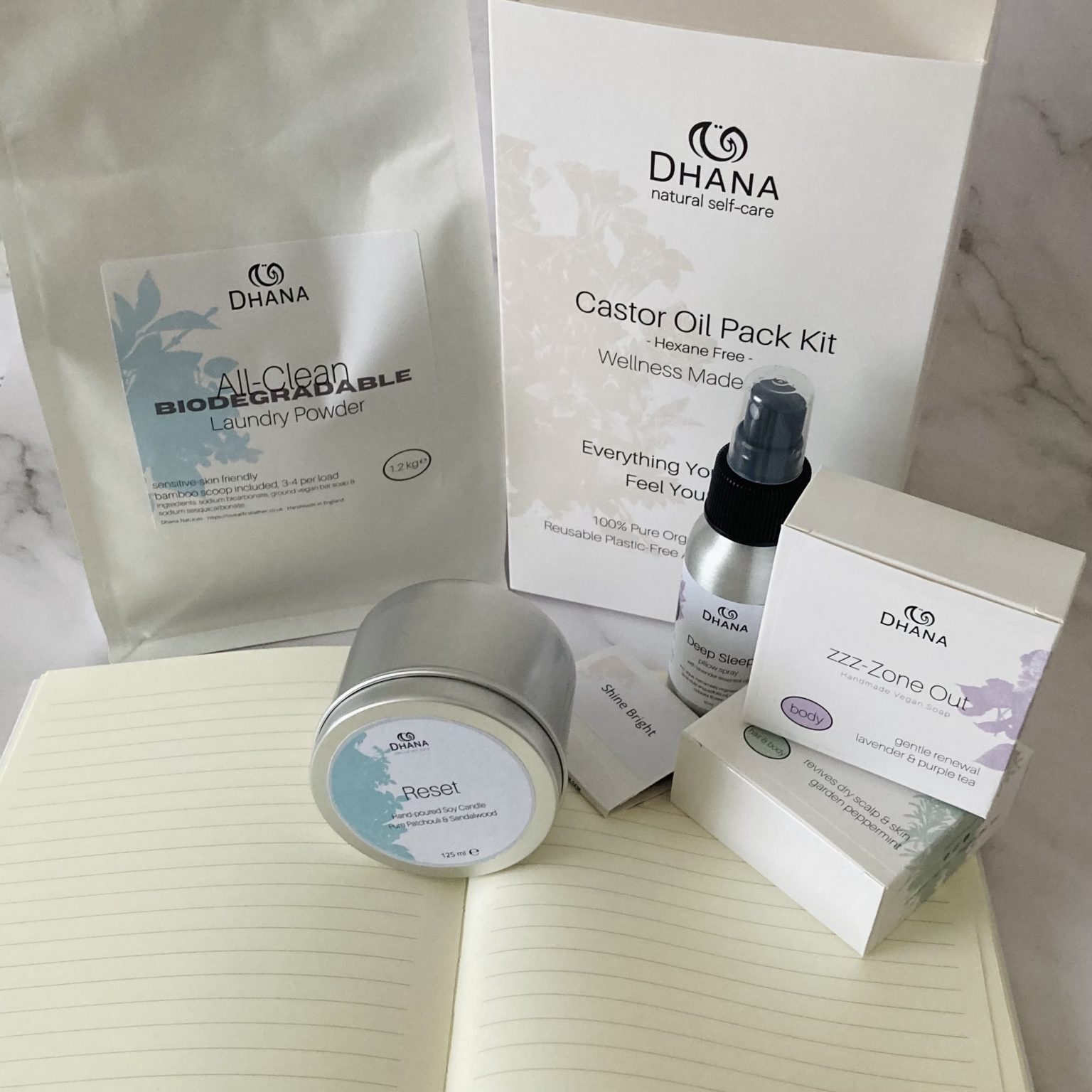 Ultimate selfcare gift (white packaged self-care items: true your journey journal, castor oil pack kit, soaps, bag of laundry powder, pillow spray and candle in tin) on marble background