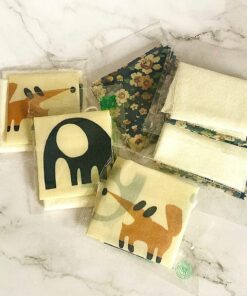 Children's Castor Oil Pack kit from Dhana Self-Care (comes with organic cotton flannel in a package with whimsical oil protectors.) Here are 6 packages with orange fox, black elephant, multi-coloured flowers and a plain flannel that each contains. All on white marble background.