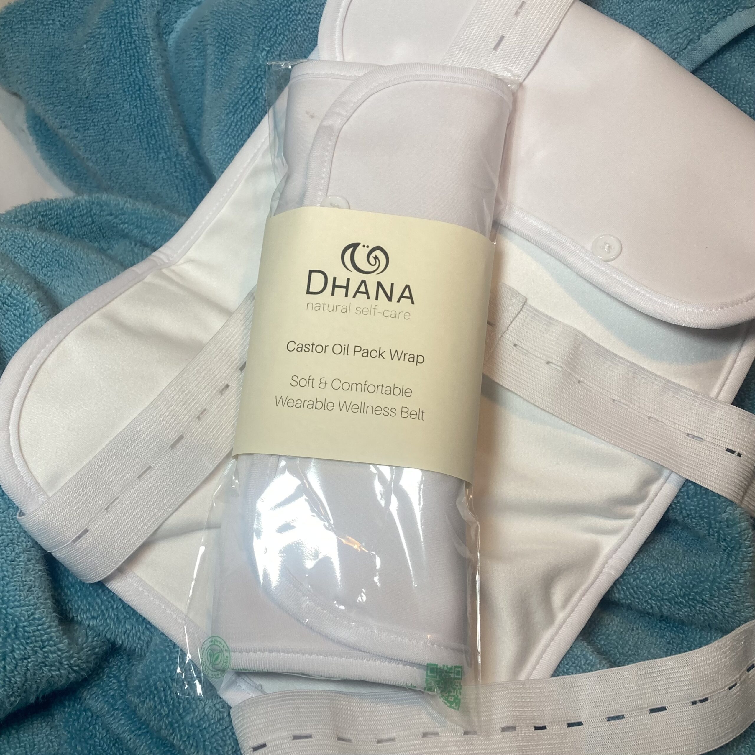 Dhana Self-Care Castor Oil Wrap in package on top of one laid out on a turquoise towel